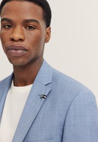 Hellblaues Sakko aus strukturiertem Stoff mit dezentem Glanz. Dekorativer Pin am Revers mit drei farbigen Streifen.