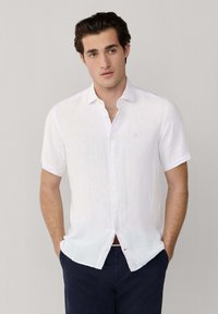 Joven con camisa blanca de manga corta abotonada y pantalones azul marino, de pie con las manos en los bolsillos contra un fondo claro y liso.