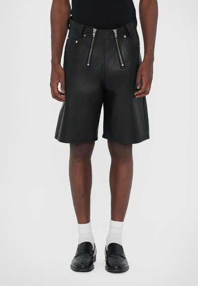 Shorts en cuir noir avec deux zips en argent à l'avant, une taille structurée et une coupe large, associés à des chaussures noires et des chaussettes blanches.