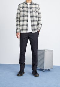 Uomo che indossa una camicia a quadri bianco e nero sopra una t-shirt bianca, pantaloni neri e stivali neri, in piedi accanto a una scatola metallica su ruote.