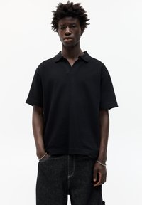 Polo nero con collo, maniche corte e vestibilità rilassata. Realizzato in tessuto morbido, indossato da un modello con jeans scuri.