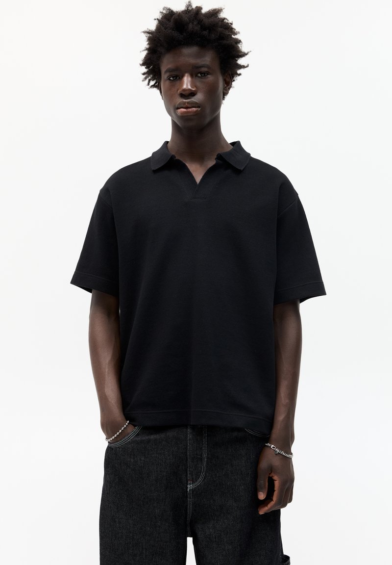 Polo nero con collo, maniche corte e vestibilità rilassata. Realizzato in tessuto morbido, indossato da un modello con jeans scuri.
