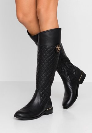 Schwarze kniehohe Lederstiefel mit Steppmuster und goldenem Emblem, getragen an überkreuzten Beinen vor weißem Hintergrund.