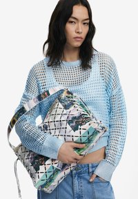 Blauer Netzpullover mit gerippten Bündchen, kombiniert mit einer großen, strukturierten Tasche, die ein bunten, abstrakten Design und diagonale Musterakzente aufweist.