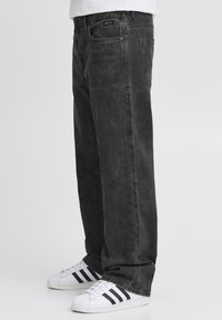 Vaqueros de mezclilla negros de corte holgado, con una textura desgastada, cinco bolsillos y herrajes metálicos. Combinados con zapatillas blancas con rayas negras.