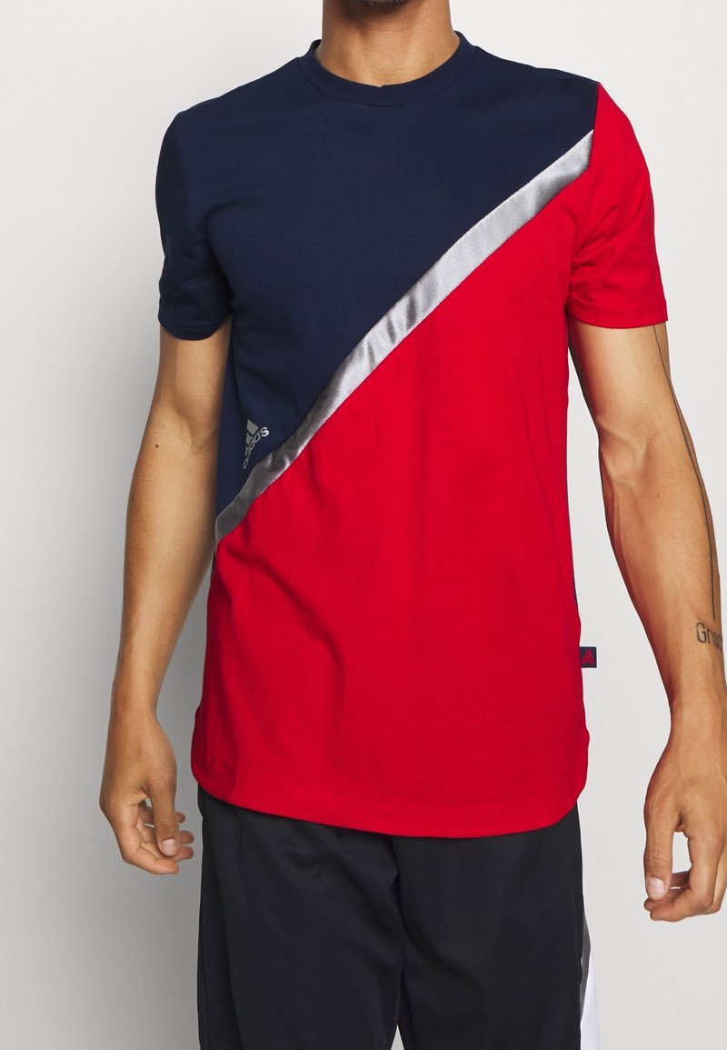T-shirt rouge et marine en coton, avec une bande diagonale argentée sur le haut de la poitrine et un logo sur le bas à gauche.