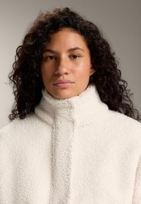 Krém színű polárdzseki magas gallérral, puha tapintású, elülső záródással. Egyszerű, letisztult kialakítású, látható minták nélkül.