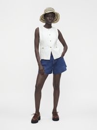 Femme portant un gilet blanc sans manches, un short en jean, des chaussures marron à plateforme et un chapeau en paille à large bord, debout avec une main dans la poche.