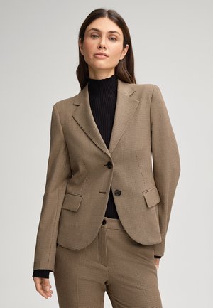 Blazer à carreaux marron et noir avec revers, deux poches avant et deux boutons. Porté par-dessus un pull col roulé noir à côtes.
