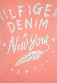 Camisa de algodón color coral con texto impreso blanco: "HILFIGER DENIM" y "Nueva York", acentuada por un gráfico de pluma y detalles de estrellas.