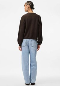 Ženska z zapletenimi lasmi v temnem puloverju in svetlo modrih širokih hlačah, stoji obrnjena stran od kamere na enostavni beli ozadju.