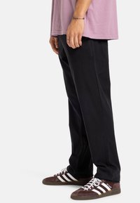 Čierne nohavice so rovnými nohavicami, nosené s voľným ružovým tričkom a hnedými teniskami so bielymi prúžkami. Minimalistický dizajn a textúra látky.