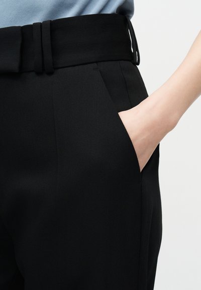 Dešinė ranka juodo diržuoto kelnio kišenėje, dėvima kartu su šviesiai mėlynu viršutiniu drabužiu, apkirptu arti juosmens ir klubų.