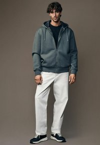 Grå zip-up hoodie med tekstureret mønster, sort t-shirt, hvide bukser og navy sneakers. Model står imod en neutral baggrund.