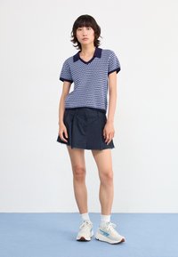 Navy and white polka dot knit polo shirt with a v-neck and navy collar, paired with a navy skirt featuring subtle pleats and frayed hem.