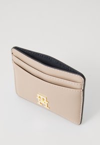 Beige leather cardholder with four slots, black interior, and gold logo accent. Smooth texture and rectangular shape.