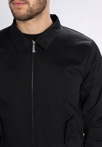 Giacca nera con colletto e chiusura con zip. Presenta tasche laterali e una texture di tessuto liscia. Design semplice e minimalista.