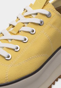 Κίτρινο πάνινο αθλητικό παπούτσι με λευκά κορδόνια που περνούν μέσα από μεταλλικές τρύπες και παχύ μπεζ καουτσούκ σόλα με υφή αντιολισθητικής επιφάνειας.