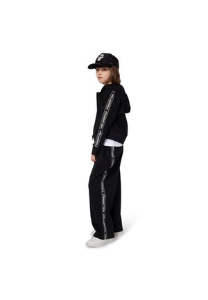 Schwarzer Kapuzenpullover mit weißem Text an den Ärmeln, kombiniert mit schwarzen weiten Hosen mit weißem Seitenstreifen. Abgerundet wird das Outfit mit einer schwarzen Mütze und weißen Schuhen.