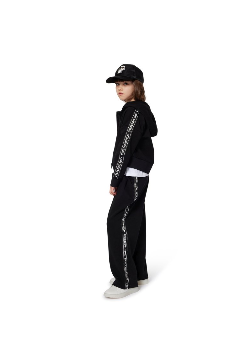 Moletom preto com texto branco nas mangas, combinado com calças largas pretas com laterais brancas. Complementa com um boné preto e sapatos brancos.
