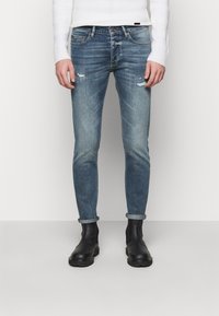 Slim-fit vybledlé modré džíny s distress efektem, ohrnutými lemy a pěti kapsami, stylizované s černými kotníkovými botami.