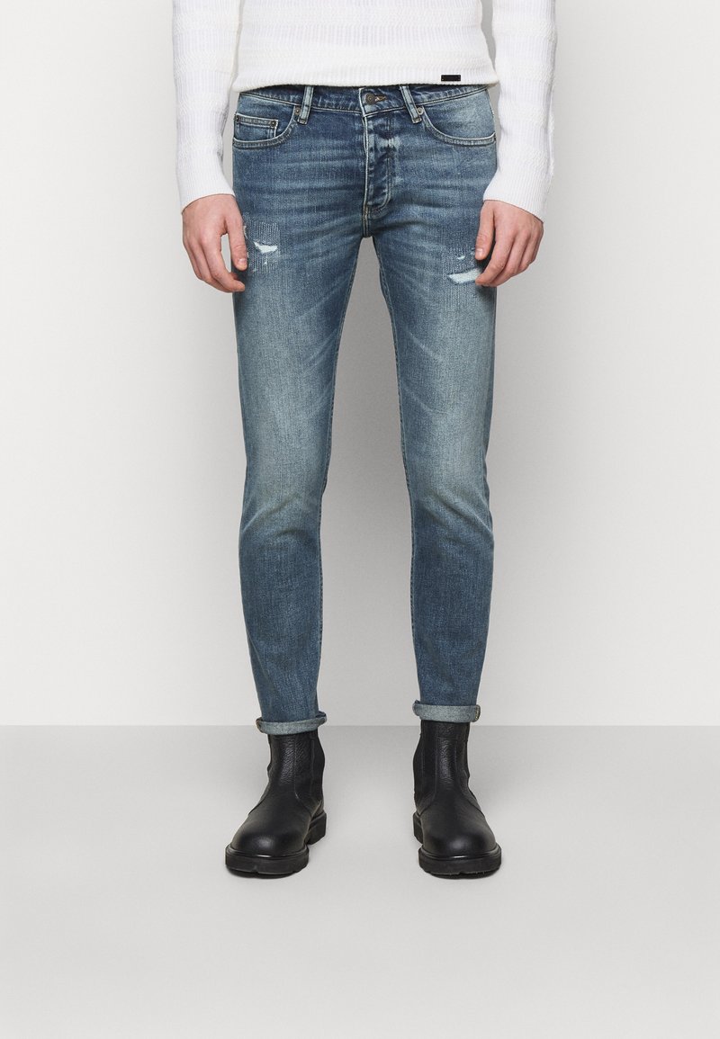 Slim-fit vybledlé modré džíny s distress efektem, ohrnutými lemy a pěti kapsami, stylizované s černými kotníkovými botami.