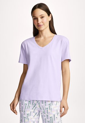 Woman with long brown hair wearing a light purple V-neck short-sleeve shirt and floral pajama pants against a plain background.