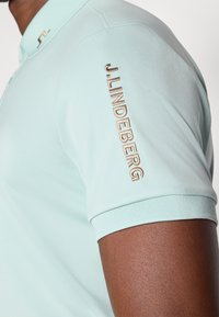Polo de cor verde menta claro feito de tecido suave, apresentando um colarinho canelado e o logótipo "J.LINDEBERG" bordado na manga em fio dourado.