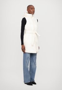 White quilted vest with a high collar and belt, worn over a black long-sleeve top. Light blue wide-leg jeans with logo detailing. Black shoes.