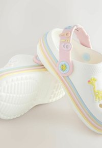 Hvide træsko med pastelfarvede regnbuestriber og gummidyrcharme. Har rillede såler, åben hælkappe og diskrete perforeringer.