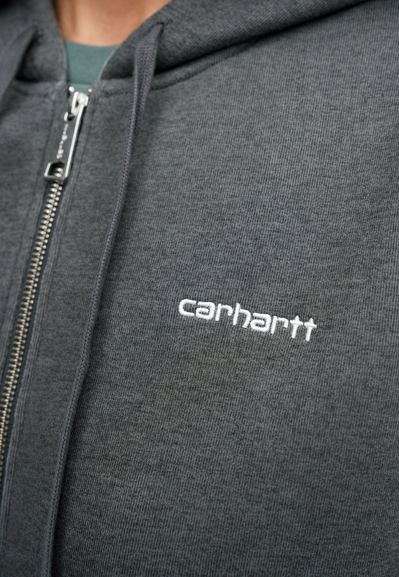 Grå zip-up hoodie lavet af tekstureret stof. Har en sølvfarvet lynlås og et broderet hvidt "carhartt" logo foran.