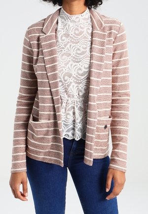 Blazer a righe in marrone chiaro e crema, con colletto sartoriale, due tasche frontali e dettaglio con bottoni. Indossato sopra un top in pizzo.