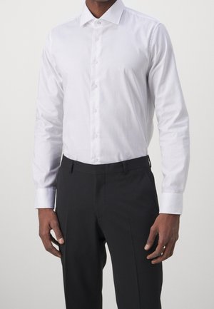 Mann trägt ein eng anliegendes weißes Hemd mit langen Ärmeln, das in schwarze, maßgeschneiderte Anzughose gesteckt ist, vor einem schlichten Hintergrund.
