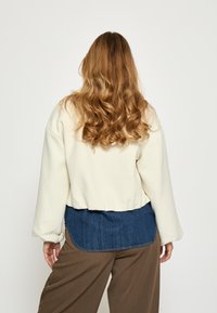 Geknipt crèmekleurig sweater met een gestructureerde stof, blauwe denim onderpaneel en een losse pasvorm. Gecombineerd met brede bruine broeken.