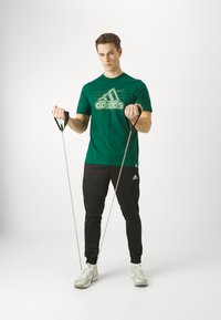 Camiseta de algodón verde con logo blanco de Adidas. Pantalones de chándal negros. Sosteniendo bandas de resistencia con mangos negros. Zapatillas blancas. Fondo sencillo.