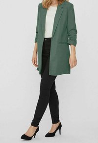 Blazer vert à la coupe structurée, manches longues retroussées, un bouton unique et deux poches latérales, associé à un jean skinny noir et des talons.