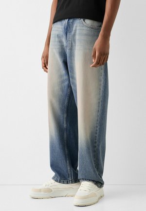 Light-wash denim jeans with a relaxed fit, featuring a faded gradient from blue to lighter hues, styled with white sneakers.