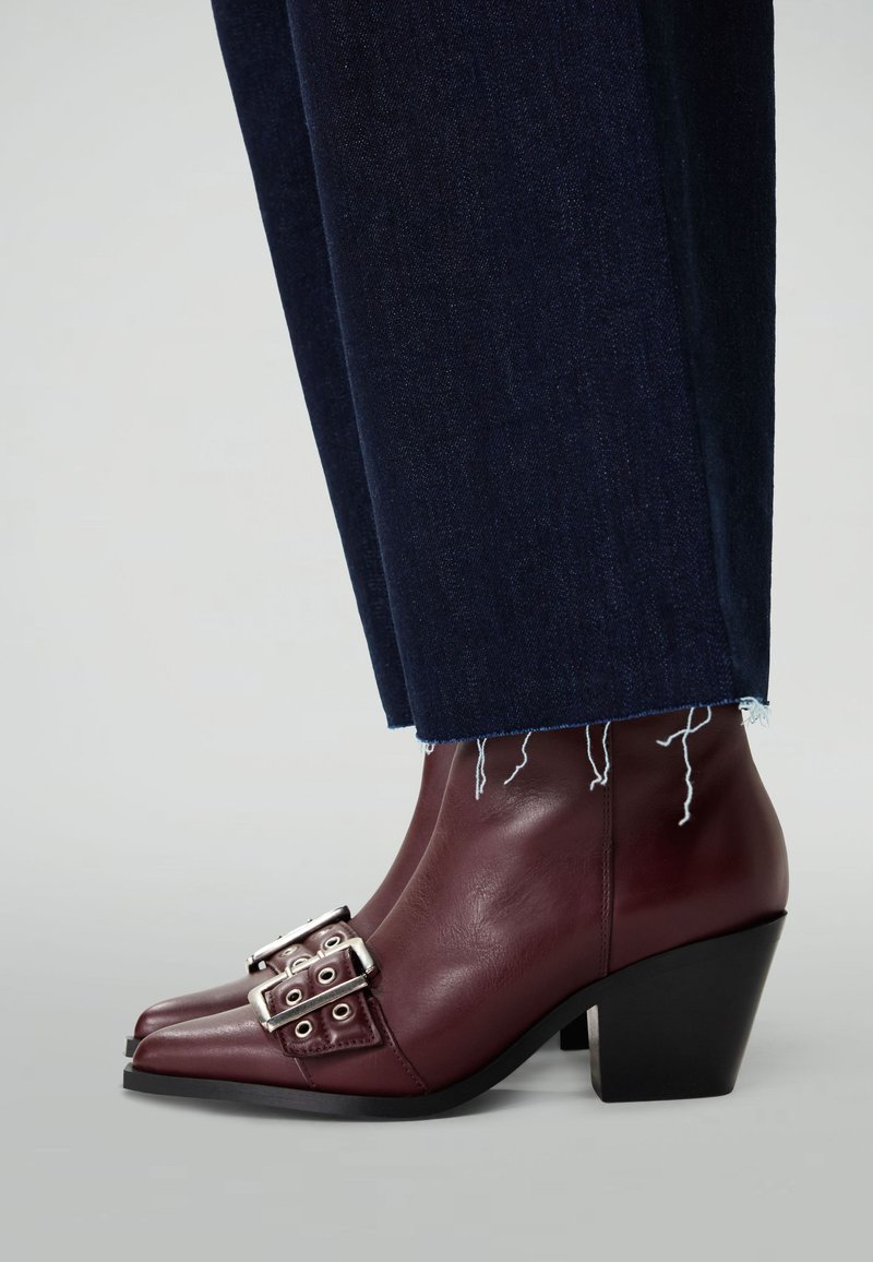 Burgundy leather ankle boots feature a square toe and stacked heel, accented with silver buckles and eyelets. Worn with dark blue denim pants.