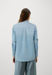 Svijetloplava traper košulja s klasičnom kragnom, dugmadi na manžetama i suptilnim naborom na leđima. Mekana tekstura, duži rukavi i zaobljen rub.