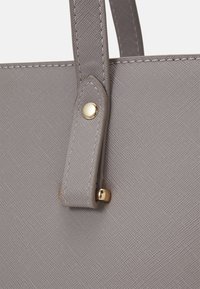 Bolso tote sintético gris con superficie texturizada, que presenta un detalle de botón en tono dorado en la correa y costuras a lo largo de los bordes.