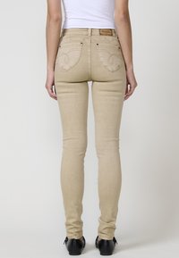 Jeans ajustados beige con una textura suave, que presentan costuras decorativas en los bolsillos traseros y un parche con el logo en la cintura.