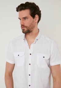 Camisa de botones de manga corta blanca con una tela texturizada, dos bolsillos en el pecho y botones marrón oscuro. Diseño de cuello clásico.