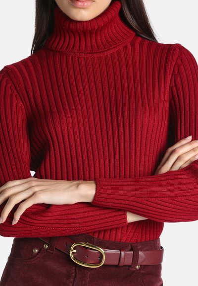Femme portant un pull à col roulé côtelé rouge, bras croisés, associé à un pantalon rouge foncé et une ceinture rouge avec une boucle ovale dorée.