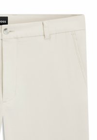 Nærbillede af beige bukser med bæltestropper, forreste knap og sidelommer, der viser fin syning og stoftekstur.