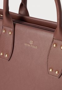 Bruine synthetische leren handtas met gouden details, voorzien van gestructureerde handvatten, een ritssluiting en het "ANNA FIELD" logo gegraveerd op de voorkant.