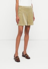 Shorts in corduroy verde oliva chiaro con design a pieghe sul davanti. Presentano una cintura e tasche laterali, abbinati a mocassini marroni con tacco.