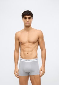 Mladý muž s kučeravými vlasmi, oblečený v svetlo šedých boxerkách Calvin Klein, stojí na jednoduchom svetlom pozadí, čelom k divákovi.