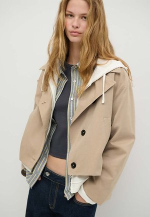 Jeune femme aux longs cheveux blonds porte des vêtements décontractés superposés comprenant une veste beige, une chemise rayée, un top court foncé et un jean sombre, les mains dans les poches.