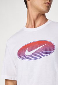 Hvit t-skjorte med kort erme som har et stort ovalt grafikk med røde og lilla horisontale linjer og en hvit Nike swoosh-logo i sentrum.