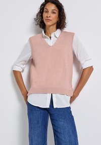 Kvinna med lockigt hår bär en ljusrosa V-ringad stickad väst över en vit skjorta med uppvikta ärmar och blå jeans framför en vit vägg.